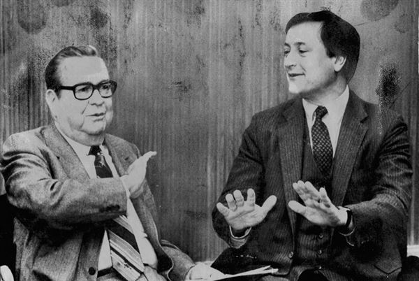 Two men sitting in a TV studio speaking to each other and gesturing.