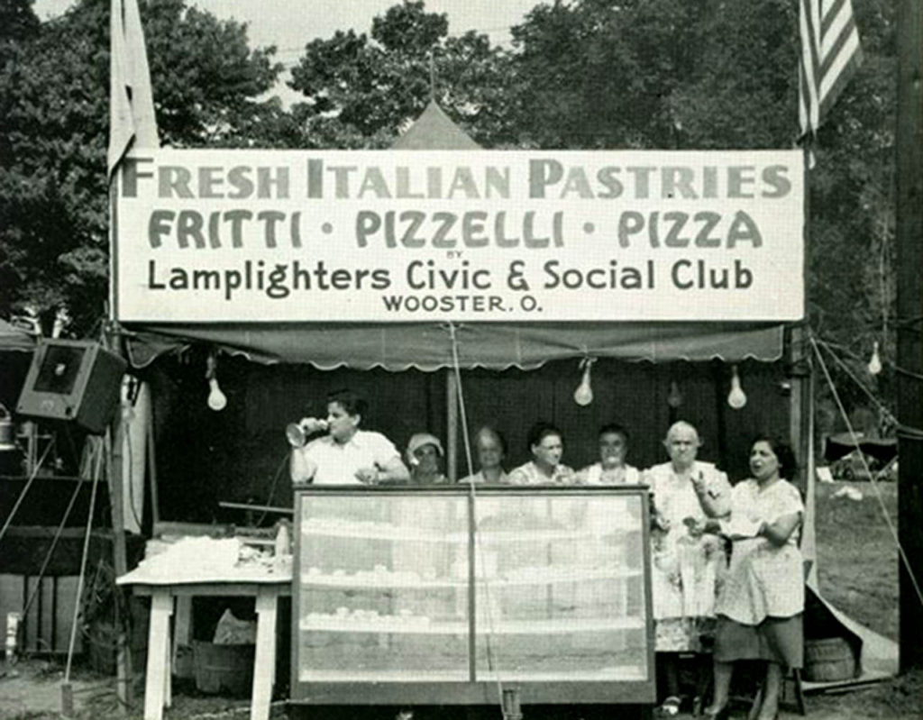 Several women standing behind a booth with a sign above them that says "Fresh Italian Pastries, Fritti, Pizzelli, Pizza, Lamplighters Civic and Social Club, Wooster, O." 