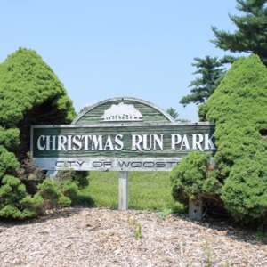 A sign that says "Christmas Run Park, City of Woster" between two bushes 