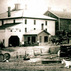 A back view of Wooster Brewing Company, which was a large farmhouse with an attached garage and a dirt back yard with a fence. 