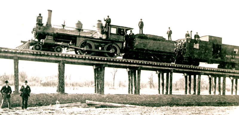 Men stand beside and on top of a train that is on elevated tracks. 