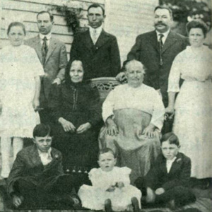 Portrait of the DiGiacomo family, with three men and two women in the back row, two older women sitting on a bench in the center, and two children and a baby sitting in the front. 