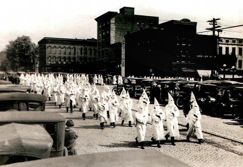 A large group of men in large white robes with white pointy hoods march down the street as people watch outside of their cars 