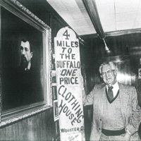 Harold Freedlander standing next to a portrait of his grandfather, holding a sign that says "4 miles to the buffalo one price clothing house, Wooster, OH" 