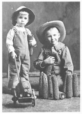 Two small children pose for a photograph, one holding a miniature pushcart and the other holding ears of corn. 