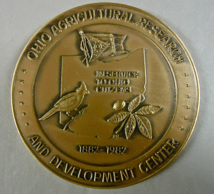 A medal commemorating OARDC's centennial. The medal says "Ohio Agricultural Research and Development Center" in the boarder, and in the center has an engraving of the state of Ohio, the state plant, the state bird, and the state flag with "In service to Ohio Citizens, 1882-1982" written on it. 