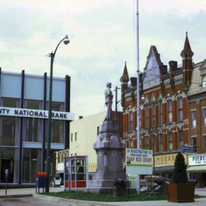 A photograph of downtown Wooster from the 1960s, that features a statue, a national bank, and businesses. 