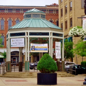 A gazebo in downtown Wooster with banners on it advertising community events, surrounded by potted and hanging flowers and trees. 