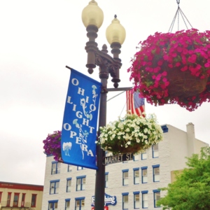 A lightpole with a banner that says "Ohio Light Opera" and hanging baskets of flowers on it. 