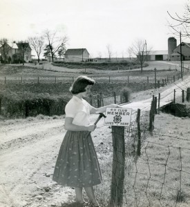 A young girl nails a sign that says "4-H Club Member Lives Here" to a fencepost, with a farm in the background, circa 1950s. 